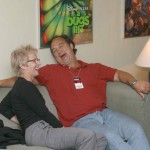 Photo of Tracy Newman and Jim Belushi seated on a couch sharing a laugh.