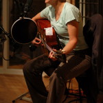 Photo of Tracy Newman seated with guitar singing into studio microphone while recording tracks in Nashville, 2009.